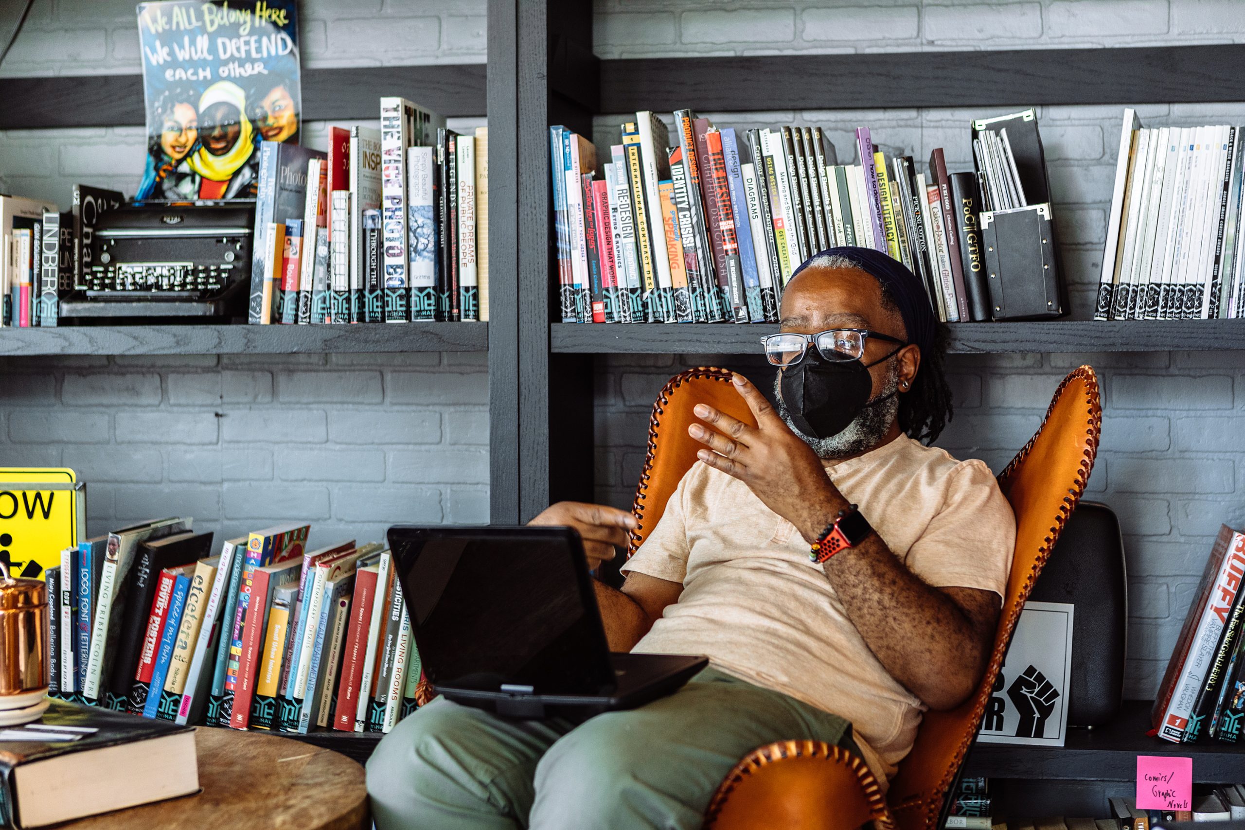 Black man in casual clothing and wearing a face mask is sitting in a chair in front of bookshelves with an open laptop on his knee.