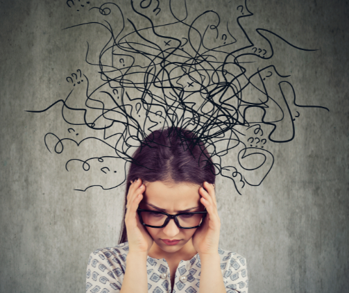 A woman wearing glasses stands in-front of a wall. Her hands are on her face and she looks upset. In the background, coming from her head are illustrated messy squiggles and question marks.