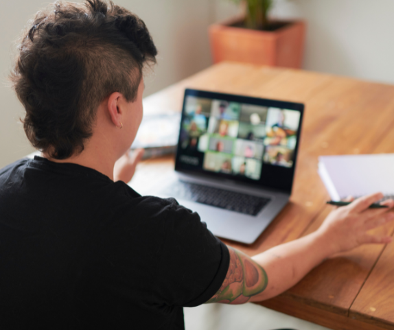 A person in a black t-shirt faces away from the camera toward a laptop screen that shows a gallery of people online together.