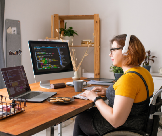 A woman in a wheelchair working at home. There is a laptop and a monitor on the desk.