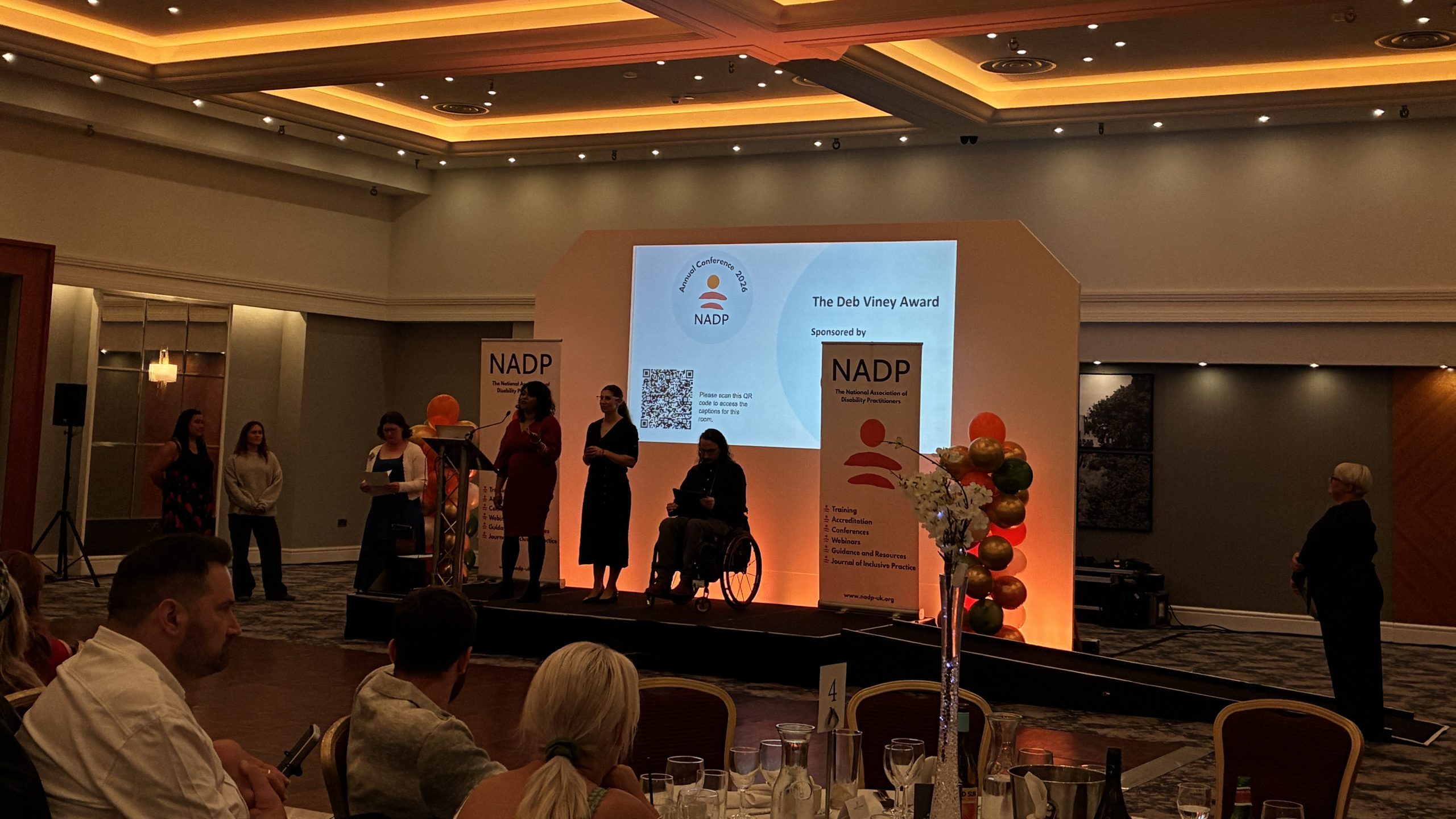 A photo from the audience of the 'Deb Viney' award being presented on-stage by two TWIP staff. They are joined two people and NADP branded banners are on-stage also.