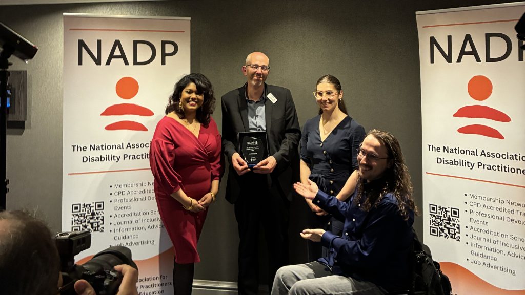 A 'behind the scenes' image of a photoshoot in-front of NADP branded banners. There are two women from TWIP and two men, one of which is in a wheelchair.