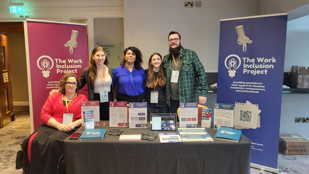 Members of The Work Inclusion Project team pose behind their exhibition table. Behind them are two TWIP branded banners.